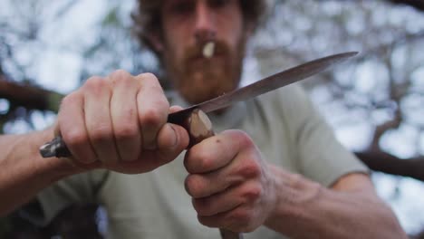 caucasian male survivalist cutting point in stick to make fire drill at camp in wilderness