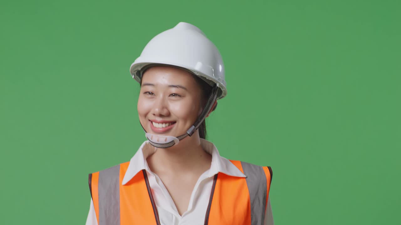 안전 헬을 입은 아시아 여성 엔지니어가 녹색 스크린 배경 스튜디오에서 는 동안 주변을 둘러보고 있습니다.