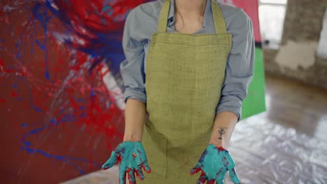 Portrait-of-a-woman-showing-hands-in-paints,-standing-next-to-her-artwork