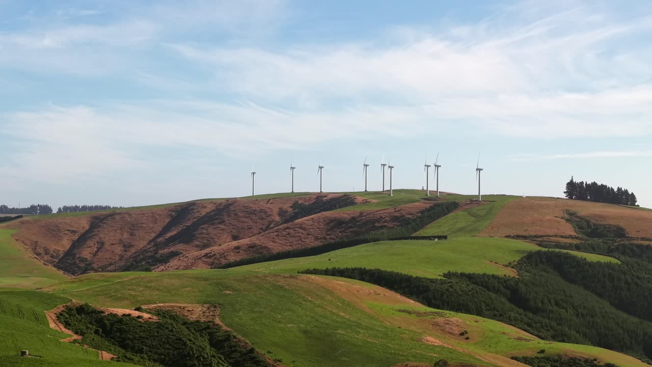 Slow drone aerial approaching and circling a wind farm atop a lush green hill, showcasing renewable energy and scenic New Zealand landscape