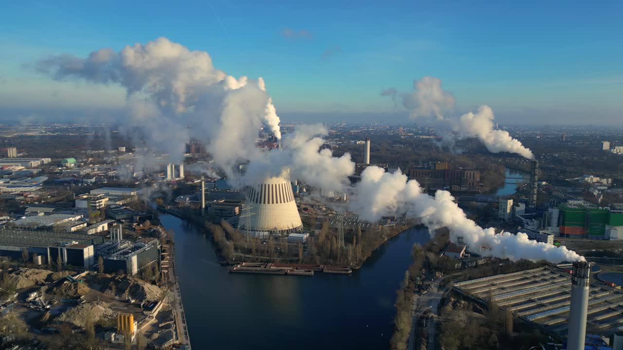 Aerial view of a thermal power plant emitting smoke on a sunny day, with a river and a city in the background. descending drone