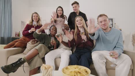 group of friends sitting on sofa, drinking wine and waving at camera at home party