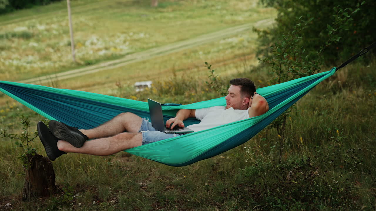 Caucasian man lies in the hammock. Relaxed freelancer works on laptop remote from the office.