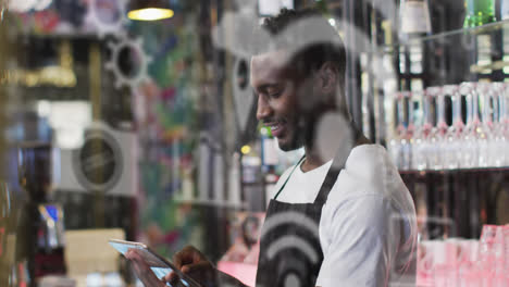 Multiple-digital-icons-floating-against-male-african-american-waiter-using-digital-tablet