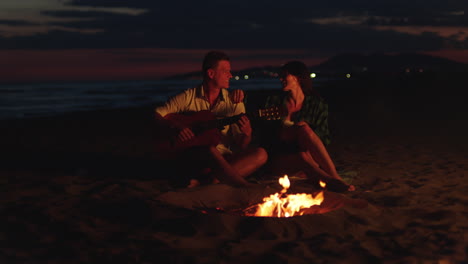 pareja tocando la guitarra junto a la fogata en la playa por la noche