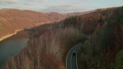 an aerial drone view shot following a car driving on a winding road alongside a lake and a water dam in late autumn