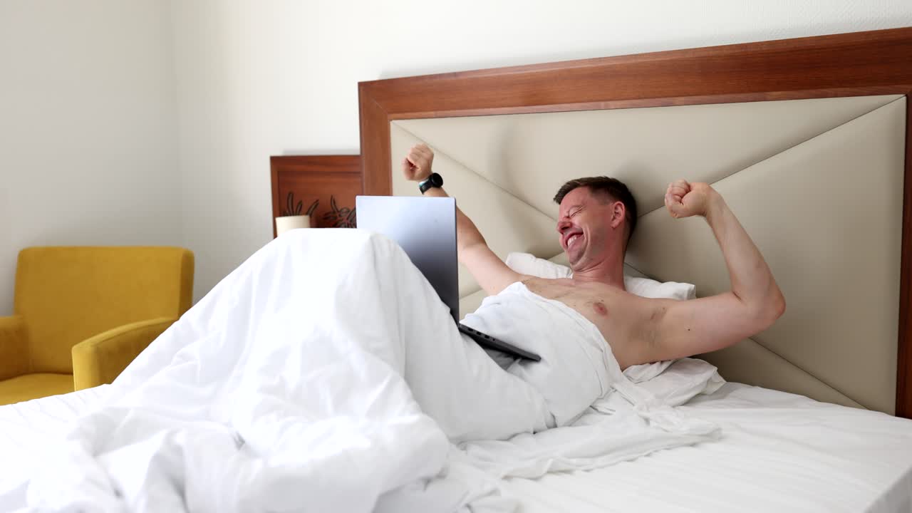 Uomo felice e bello sdraiato sul letto ride e legge un messaggio al computer.