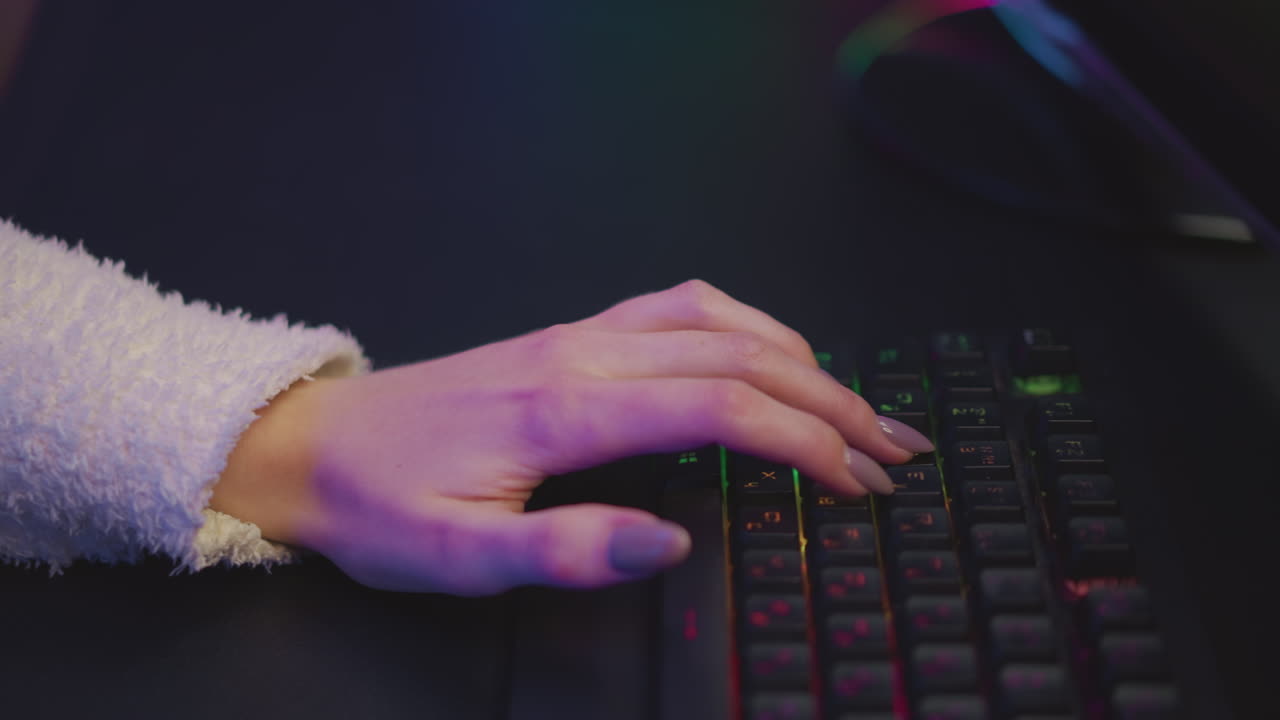 Top down view of girl hand in fluffy teddy bear sweater typing on illuminated mechanical keyboard with colorful RGB backlight, showing polished nails and concentrated typing in cool gaming atmosphere