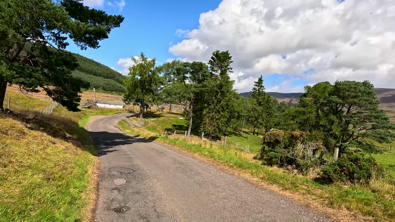 Vehicle drives along winding rural road in scenic Scottish countryside under bright daylight, dynamic camera