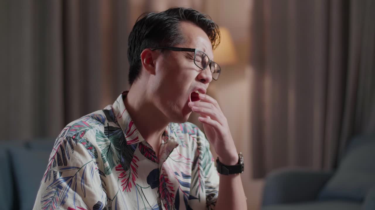 체어에 앉아 있는 피곤한 아시아인 남자가 집에 있는 카메라 에 있는 데스크에서 영상을 편집하는 동안 거리고 있다.