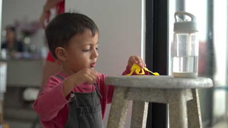 Little-latin-hispanic-boy-playing-with-a-yellow-broken-plastic-frog-toy-on-a-wooden-stool