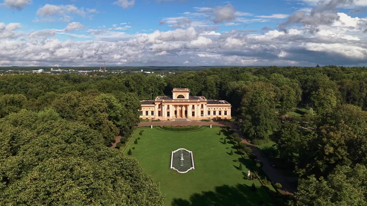 Drone video showcases Trakų Vokė Manor estate near Vilnius, Lithuania, framed by green parkland and dense trees under dramatic clouds, in crisp daylight with wide aerial composition.