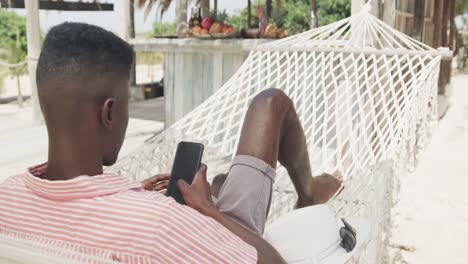 Hombre-Afroamericano-Tumbado-En-Una-Hamaca-Usando-Un-Teléfono-Inteligente-En-Una-Playa-Soleada,-Cámara-Lenta