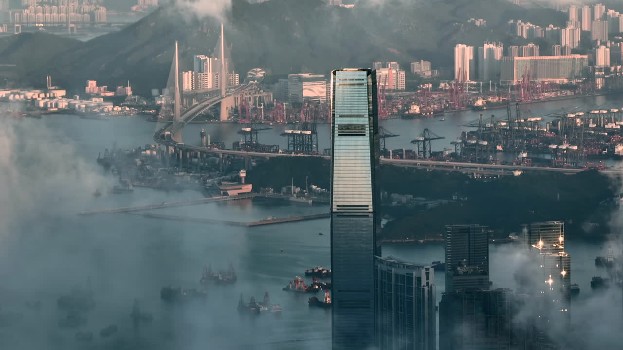 Revealing ICC skyscraper in Hongkong behind clouds with container port and stonecutters bridge in the background during an early morning. Zoomed drone panning shot