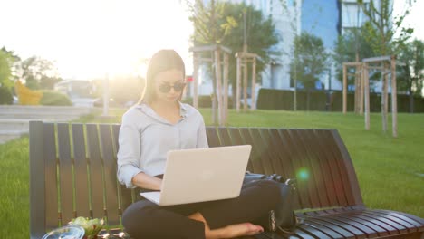 business woman is sitting on bench outdoors and using lap top for working out office and eating salad