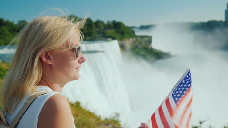 reisen in den usa - eine frau mit der flagge von amerika in der hand bewundert die niagarafälle