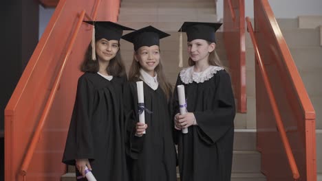 tres felices estudiantes de preescolar con toga y birrete de pie en las escaleras, sosteniendo un diploma y hablando juntas en la ceremonia de graduación