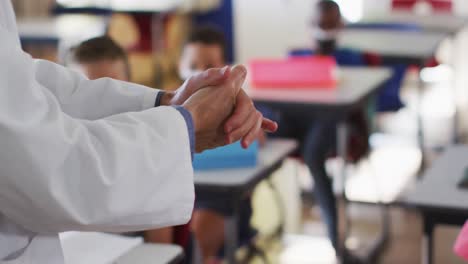 Diverse-teacher-disinfecting-hands-in-classroom,-with-schoolchildren-sitting-wearing-face-masks