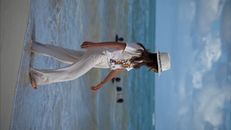 Cámara-Lenta-Vertical-De-Una-Hermosa-Mujer-Morena-Latina-Caminando-Por-La-Playa-Usando-Pantalones-De-Lino,-Un-Sombrero-Blanco-Y-Gafas-De-Sol-Amarillas-Disfrutando-Del-Mar