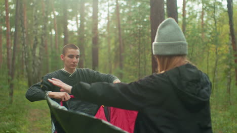 students coordinate setting up camping tent in forest, stretching tent fabric between them while surrounded by pine trees and soft natural sunlight filtering through woodland in early morning calm