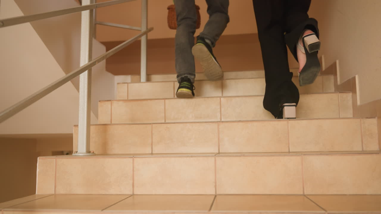 couple holding hands climbing stairs, intimate indoor staircase scene showing couple ascending tiled steps in apartment hallway, casual clothing, rear view, wicker basket, warm beige walls, soft