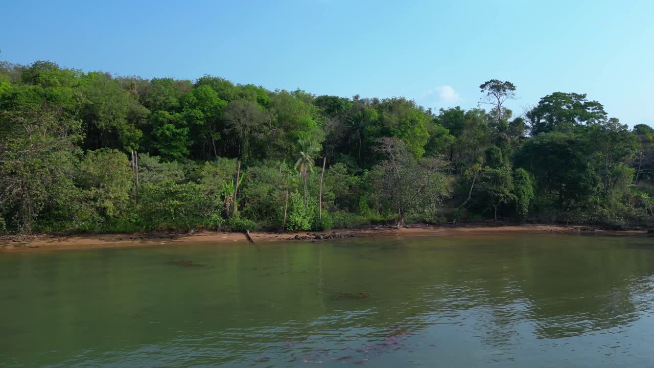 Lush vegetation covering tropical island coast meeting calm ocean water and small sandy beach. Smooth aerial view flight panorama orbit drone