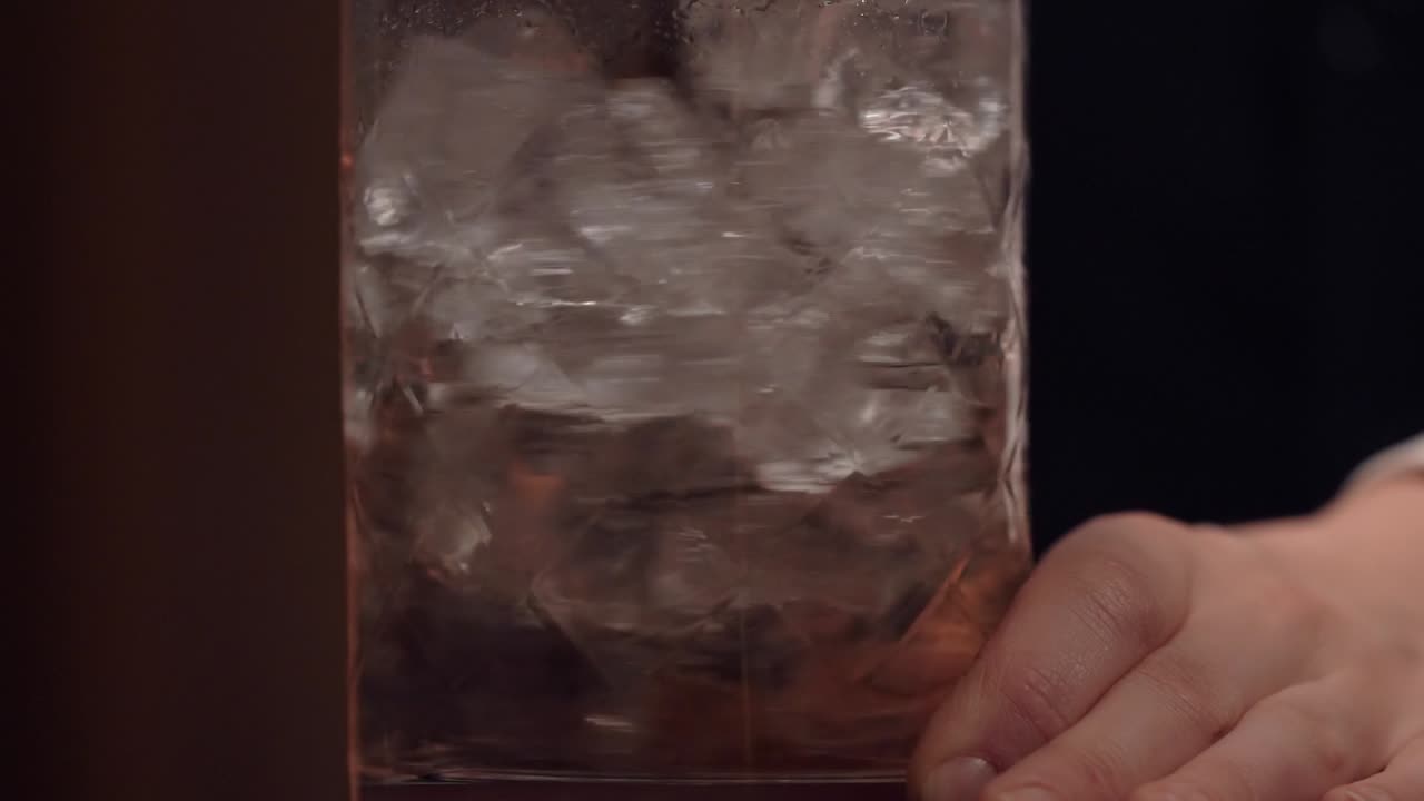 A bartender is pouring spirit and liquor into the stirring glass and start to stir along a lot of ice using a long copper bar spoon.