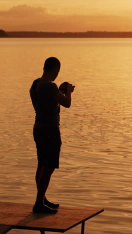 a talented photographer with a camera in his hands taking pictures of the sunset on the river on the bridge with a bench. silhouette of man with a camera on his favorite work. vertical video