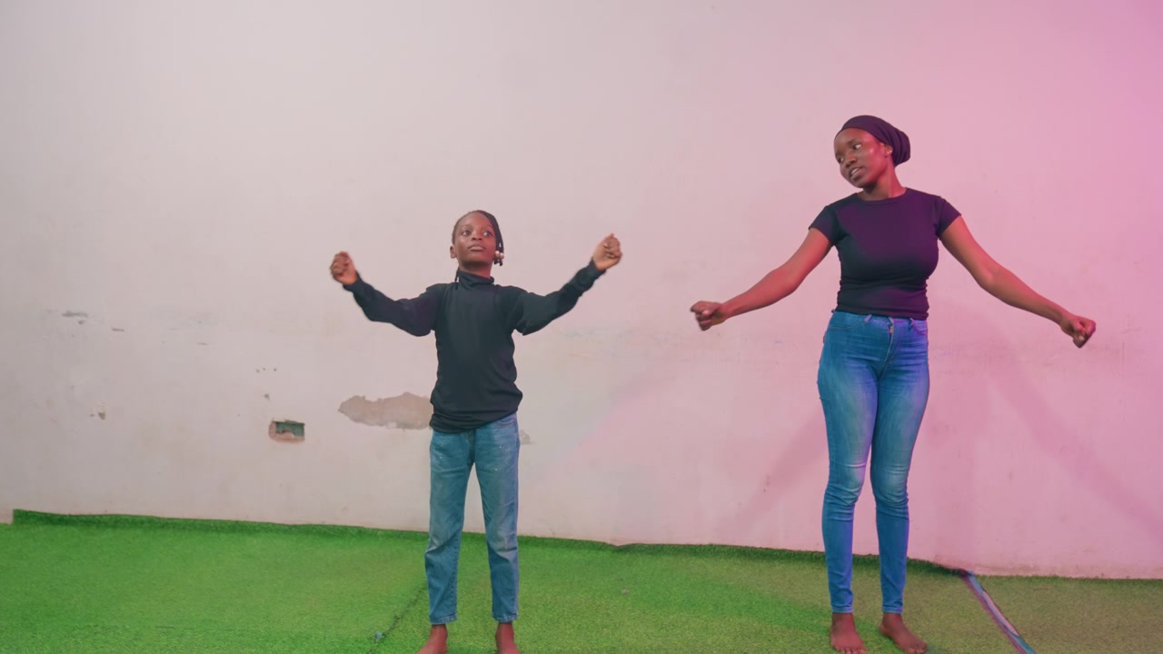 Black mother and child jump together indoor studio with pink light and green turf, mom leading joyful mimic, arms raised, casual denim outfits, upbeat playful energy and bonding moment