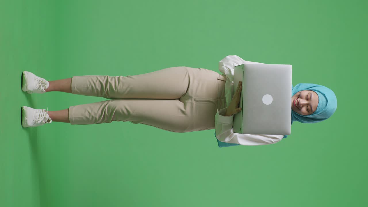 cuerpo lleno de mujer musulmana asiática sonriendo y escribiendo en una computadora portátil mientras está de pie en el estudio de fondo de la pantalla verde