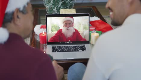 Padre-E-Hijo-Birraciales-Sonrientes-Usando-Una-Computadora-Portátil-Para-Una-Videollamada-Navideña-Con-Santa-En-La-Pantalla