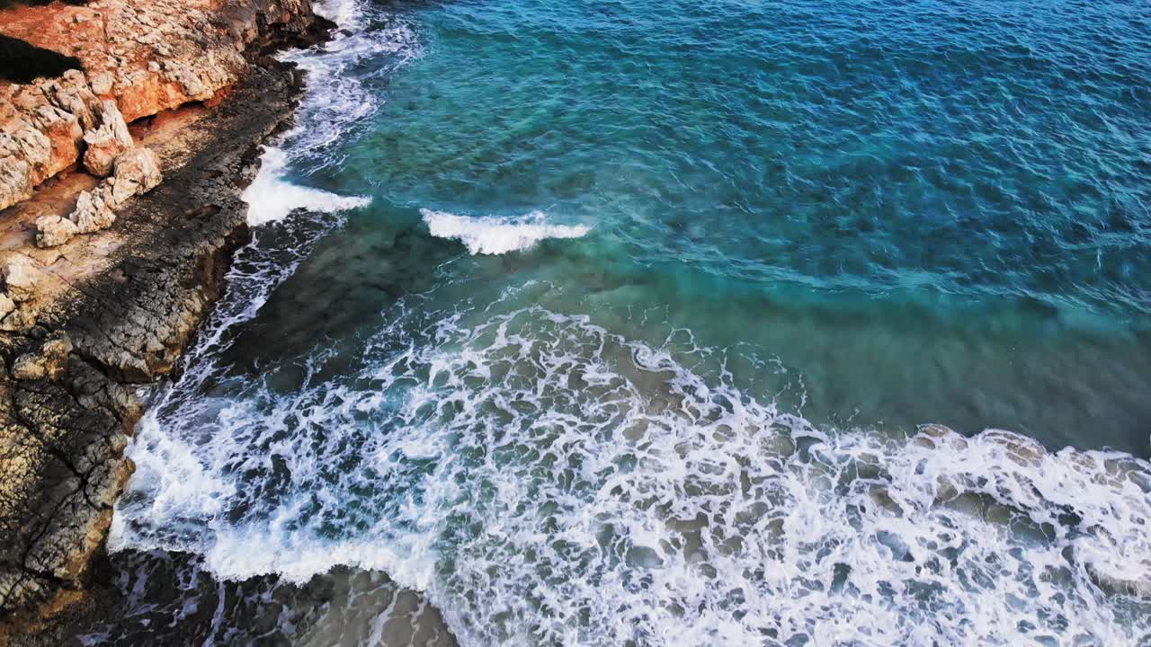 aerial drone shot of a rocky coastline in Majorca with waves crashing onto the rocks and sandy beach of the Mediterranean