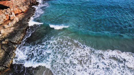 aerial drone shot of a rocky coastline in majorca with waves crashing onto the rocks and sandy beach of the mediterranean