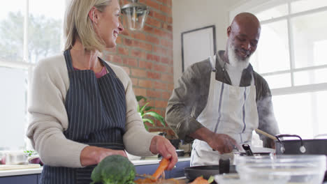 Una-Pareja-De-Ancianos-De-Raza-Mixta-Usando-Delantales-Cocinando-Comida-Juntos-En-La-Cocina-De-Casa