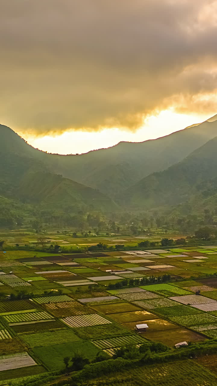 Sunset rays burst through thick clouds illuminating square crop plots in valley below, vertical aerial establishing