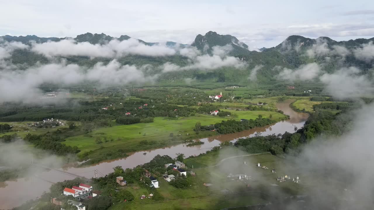 Drone terbang melewati awan yang memperlihatkan desa-desa kecil Vietnam dan sawah di sepanjang sungai berangin dengan jajaran pegunungan hijau di latar belakang di Phong Nha, Vietnam.