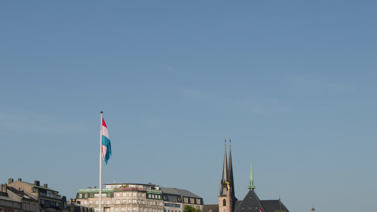 Contemporary tram glides past Luxembourg cityscape, cathedral spires, and flag in golden sunset light