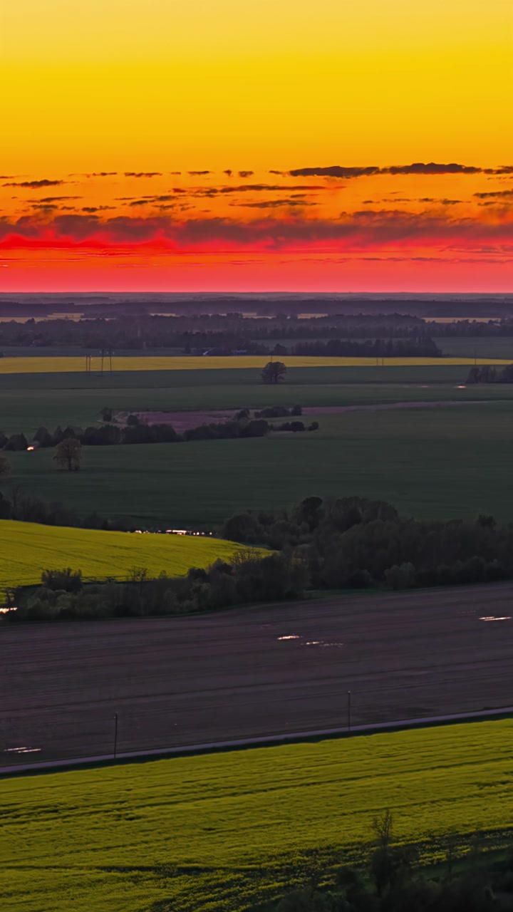 Drone hyperlapse glides over flat agricultural fields and tree clusters in Latvia beneath a blazing red and orange sunset sky at dusk
