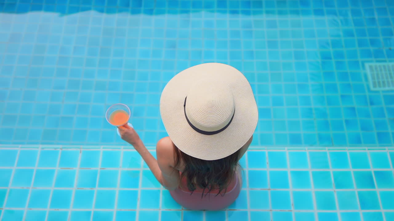 Young beautiful women wearing a swimsuit sitting on the edge of the swimming pool, enjoying the drink
