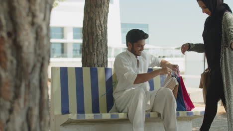 Muslim-man-sitting-on-bench,-meeting-wife-with-shopping-bags.