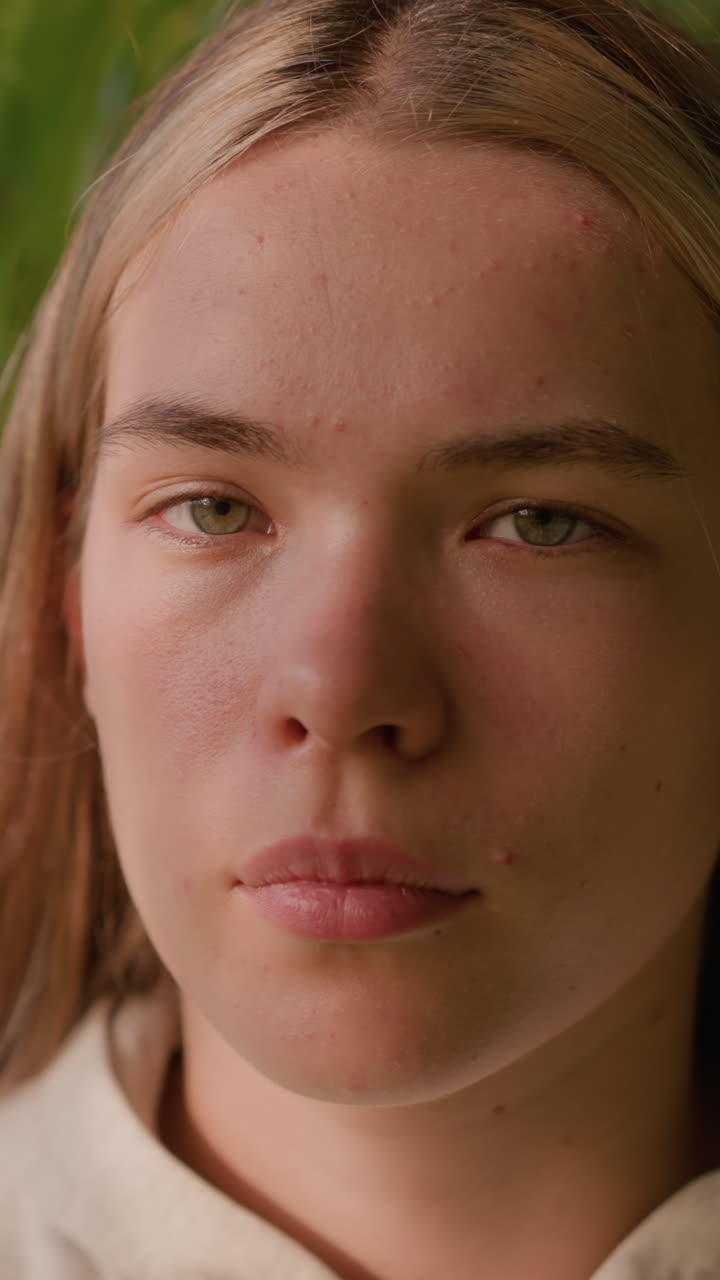 joven con expresión enfocada, de pie al aire libre con intensidad tranquila en un entorno natural, la luz del sol arroja sombras suaves en su rostro, mezclándose con la vegetación de fondo