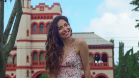 Latina-Mit-Lockigem-Haar-Lächelt,-Während-Sie-Auf-Einem-Baumstamm-Mit-Blauem-Himmel-Und-Einem-Schloss-Im-Hintergrund-Sitzt