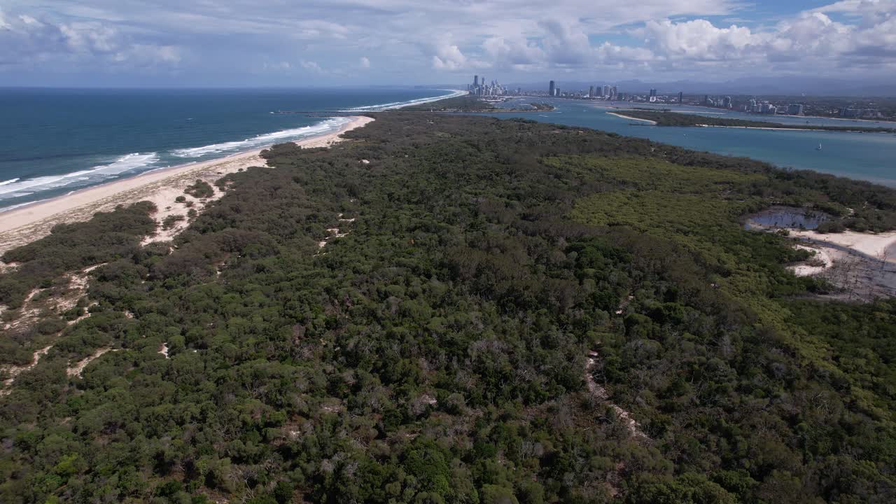Aerial View Of South Stradbroke Island With Dense Coastal Forest And Sandy Shores. Gold Coast City Skyline In Distant Background.