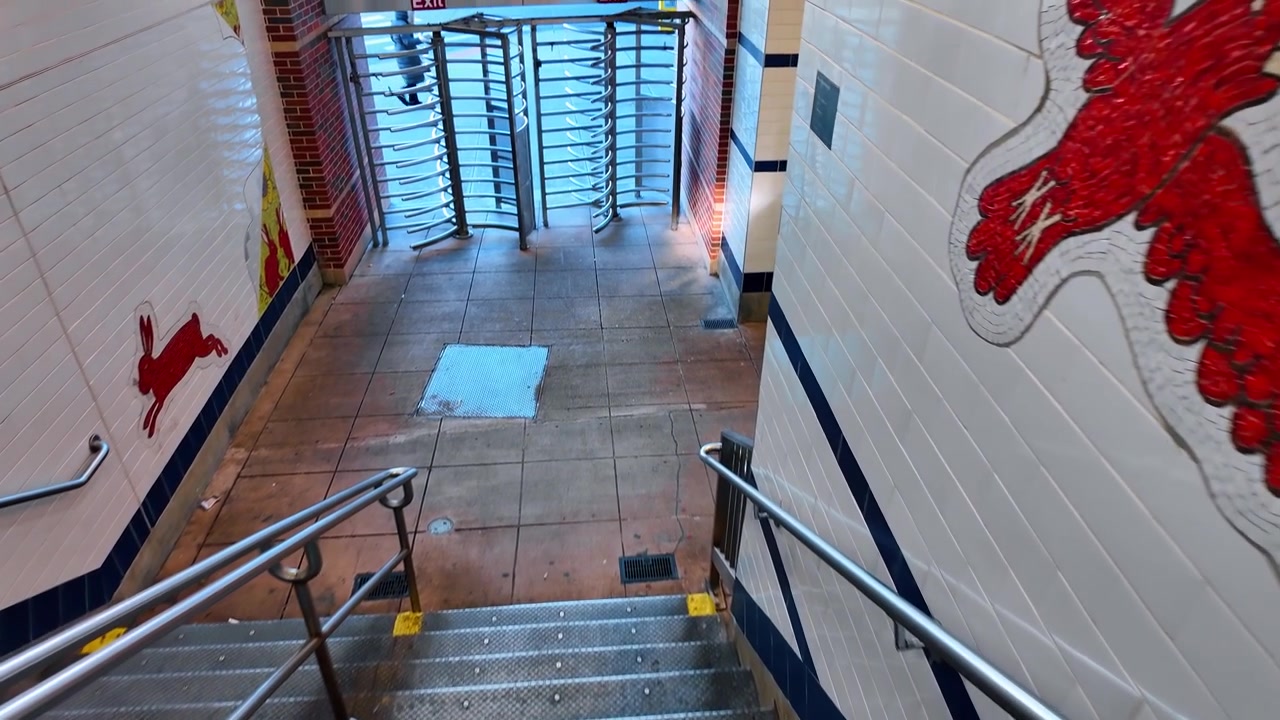 Uscita con scala e tornello all'interno della stazione della metropolitana Avenue M. Una vista dall'alto guarda giù i gradini piastrellati verso cancelli metallici, finestre a blocchi di vetro e un'opera d'arte murale con un coniglio rosso.