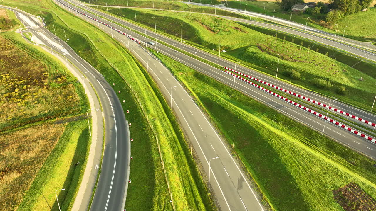 Modern highway lanes and on-ramps cut through bright green, manicured slopes. A single white car drives past red-and-white safety barriers in this sunny aerial shot