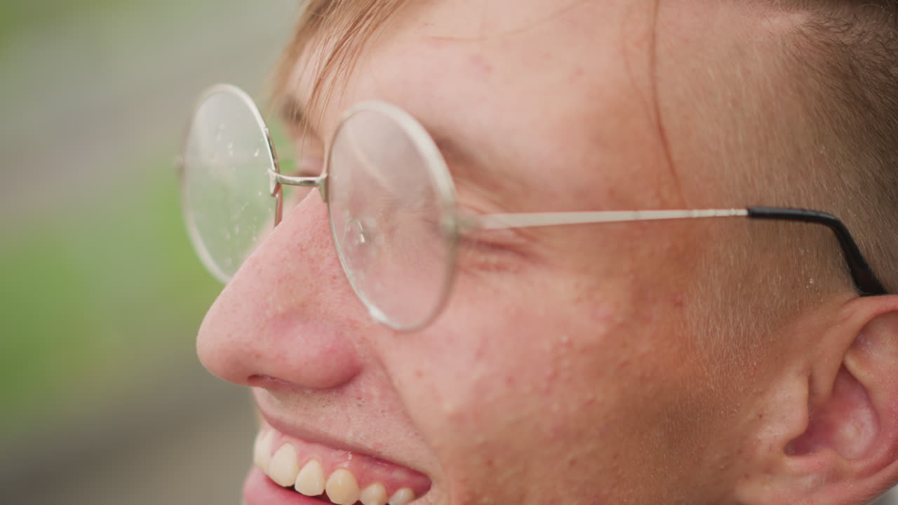 젊은 백인 남성의 옆모습, 미소를 짓고 있고, 빗방울이 비치는 젖은 둥근 안경을 쓰고, 초록색으로 흐릿하게 보이는 공원 배경, 바람에 헝클어진 머리, 자연스러운 웃음, 캐주얼 재킷, 장난기 넘치는 통근자의 활기, 역할