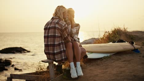 Happy-couple-a-blond-man-in-a-checkered-shirt-sits-on-a-wooden-bench-on-his-legs-sits-a-blonde-girl-in-a-checkered-shirt-and-a-hat-they-hug-and-kiss-near-their-surfboards-on-strawberries-near-the-sea-at-Sunrise-in-the-morning-in-summer