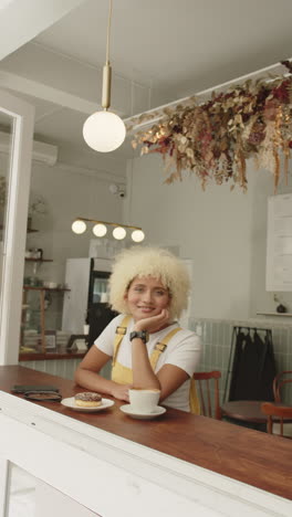 vertical video: woman in cafe smiling, sitting at counter with coffee and donut