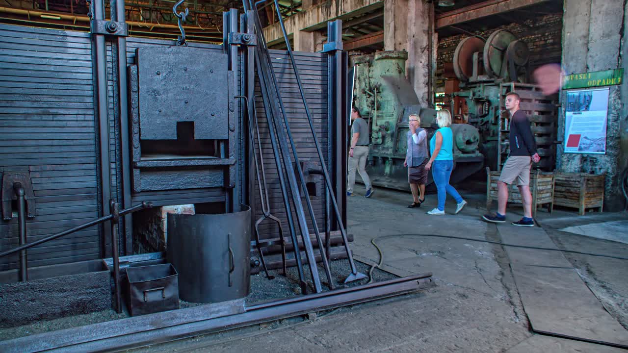 Family of four takes a tour in old steel mill with huge industrial machines. The guide explains the devices. Tracking shot