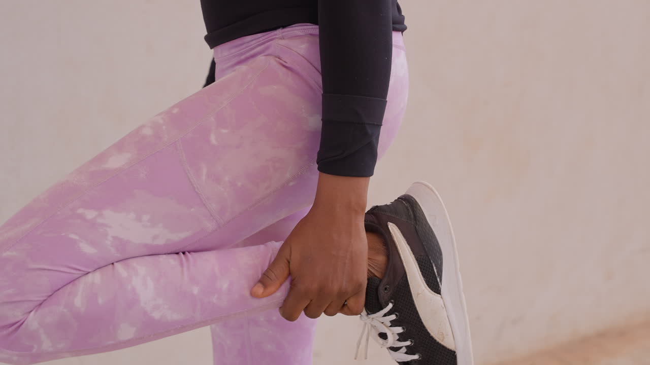 black woman stretching leg against wall in studio, wearing lavender leggings and black sneakers, focused pre workout warmup led by trainer, gentle quad hold, neutral minimal background and calm